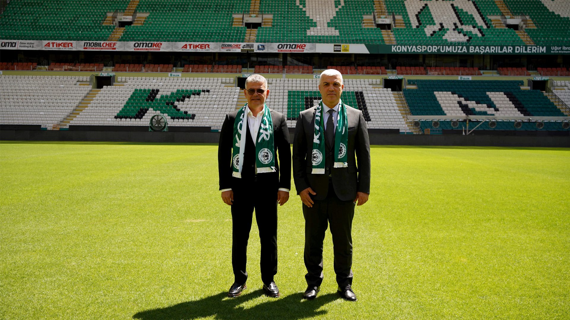 Two men in suits and green scarves stand on a football pitch in a stadium with green and white seats.