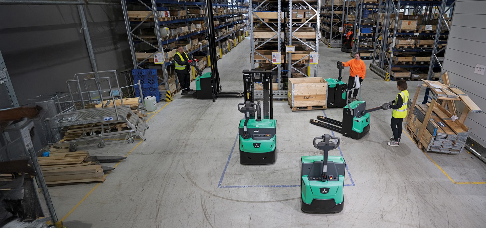Warehouse workers operate green electric pallet trucks and stackers amidst storage shelves.