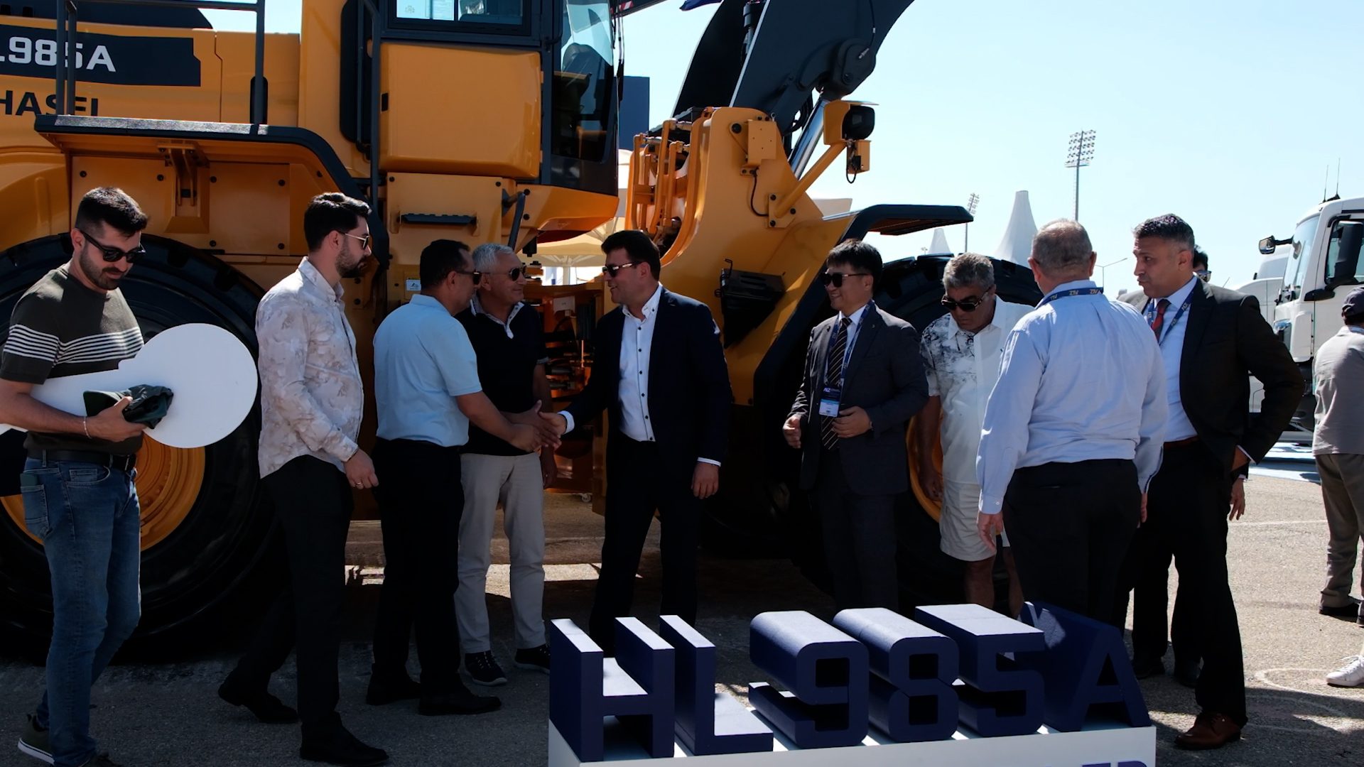 Businessmen shaking hands beside a large yellow construction loader and an 'HL985A' display.