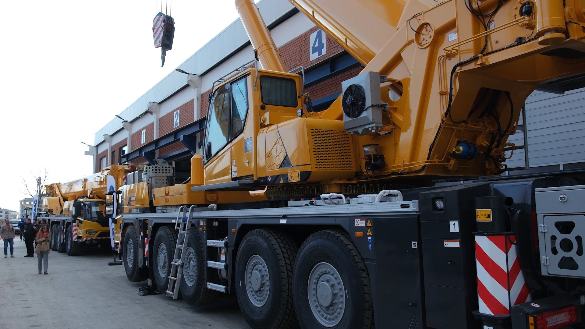A large yellow mobile crane parked outside a building, with other cranes and people in the background.
