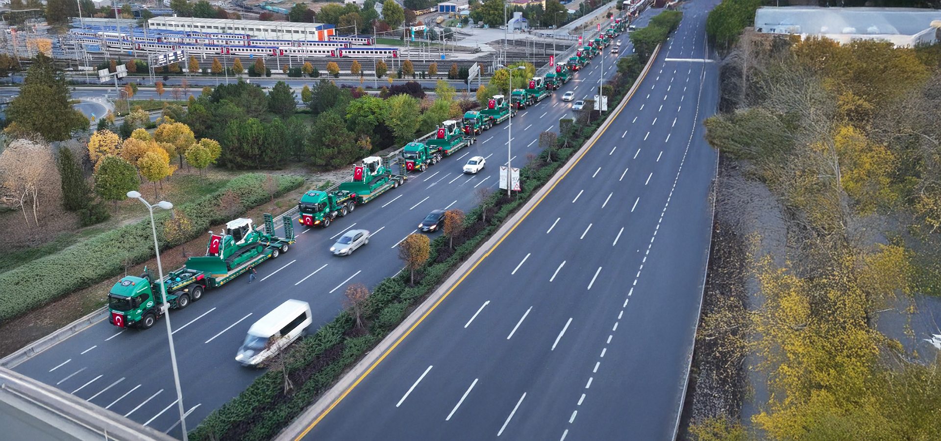Convoy of green trucks with Turkish flags carrying heavy equipment on a highway, train station in background.