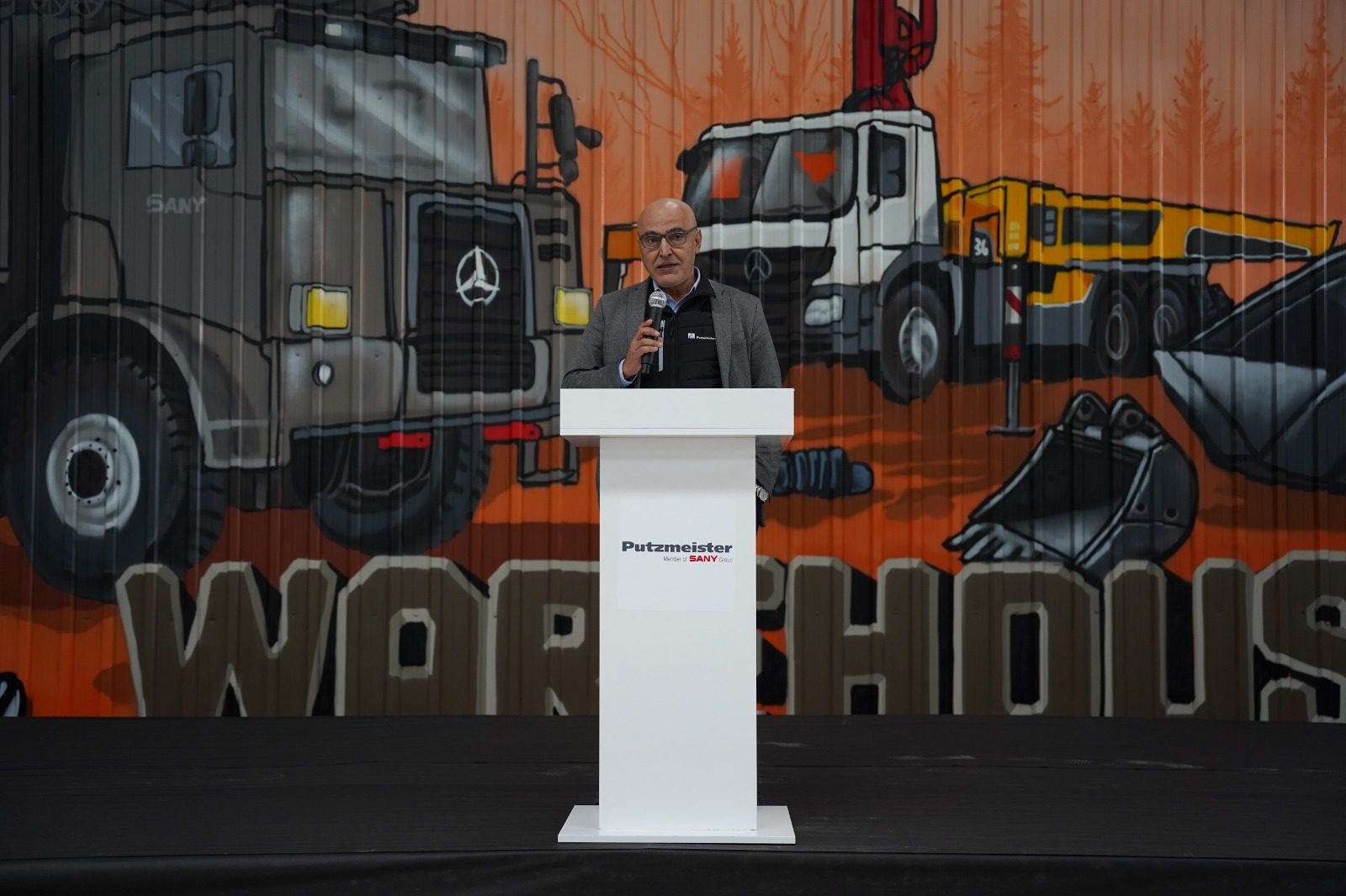 Man at podium with "Putzmeister Sany" logo, in front of a mural of construction vehicles.