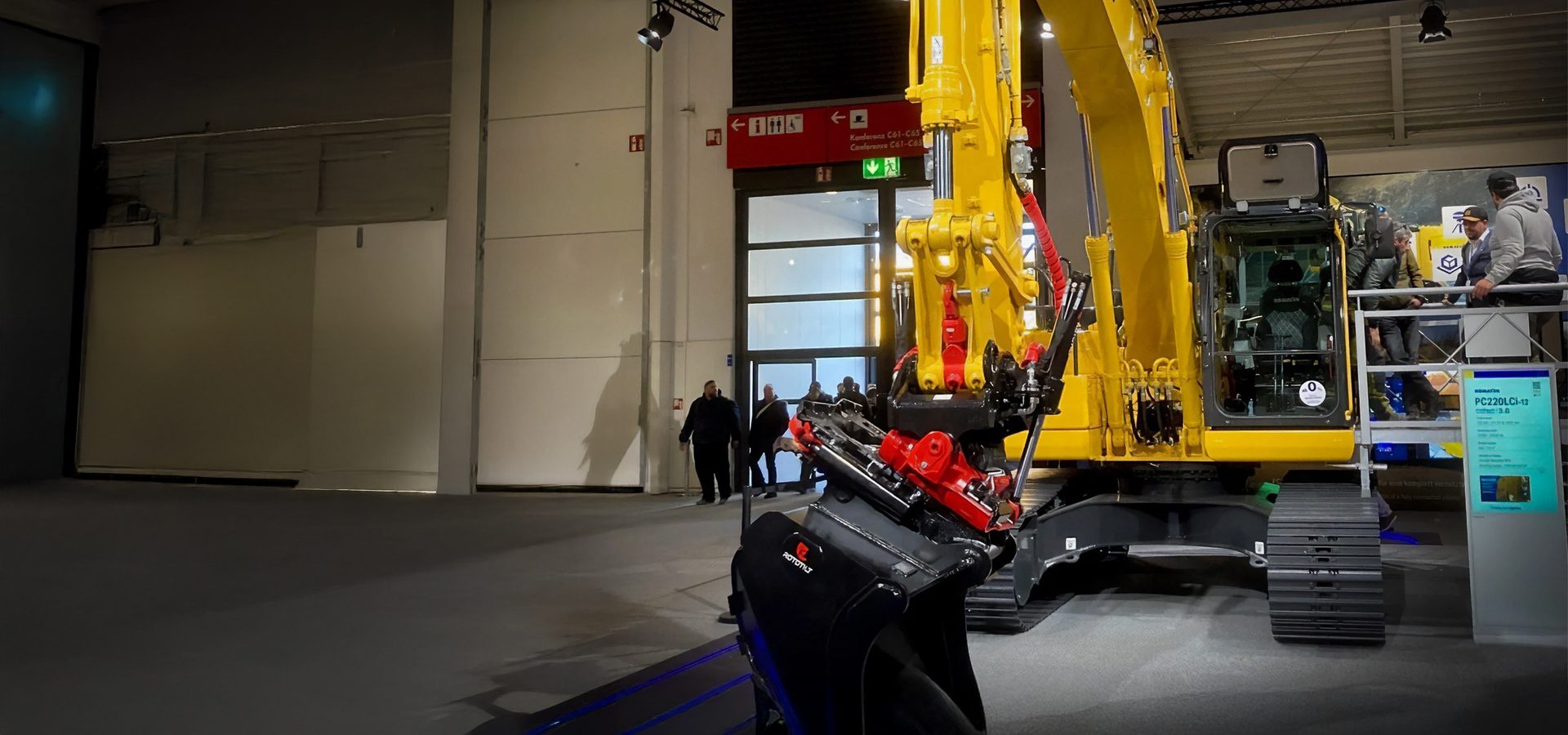 Yellow Komatsu PC220LCI-12 excavator with Rototilt attachment at an indoor exhibition.