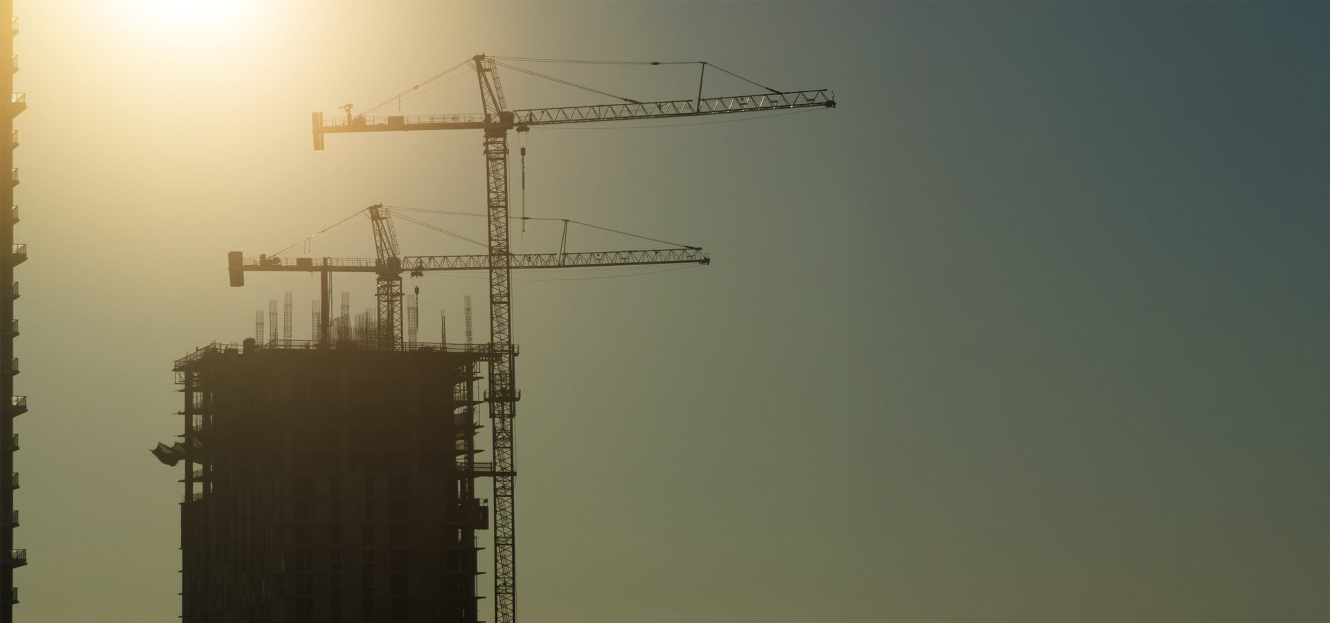 Silhouettes of construction cranes and a building against a hazy sky with a bright sun flare.