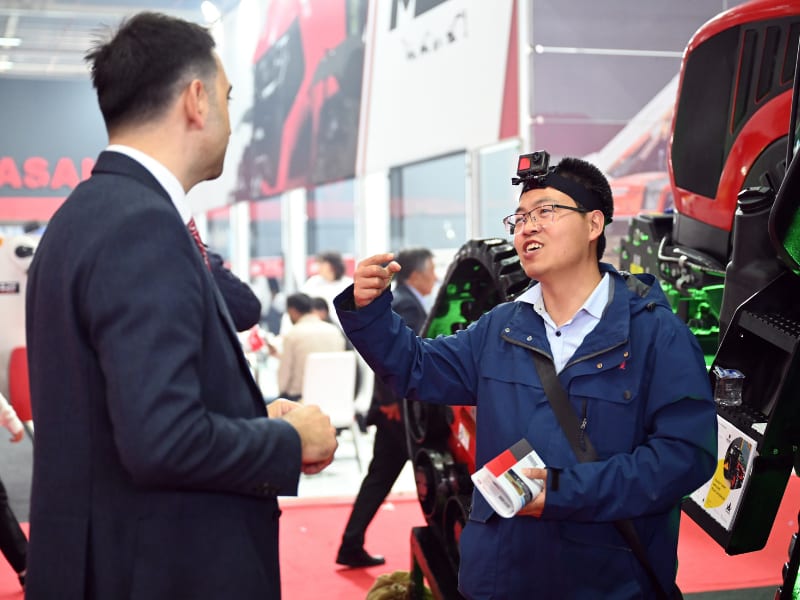 Two men discuss at an exhibition, one wearing a head-mounted camera and pointing.