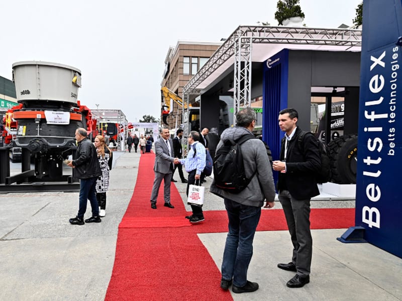 Attendees at an outdoor industrial trade show with heavy machinery and a prominent red carpet.