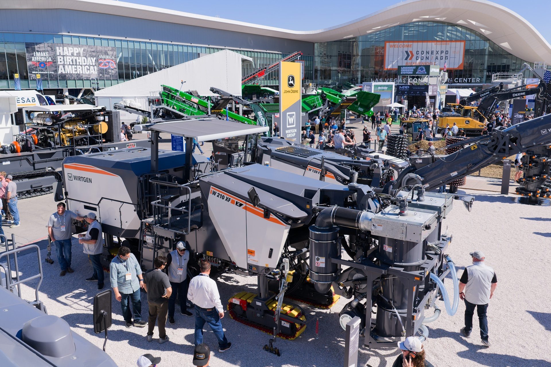 Outdoor exhibition of large construction machinery, including Wirtgen and John Deere, with many attendees at Conexpo.