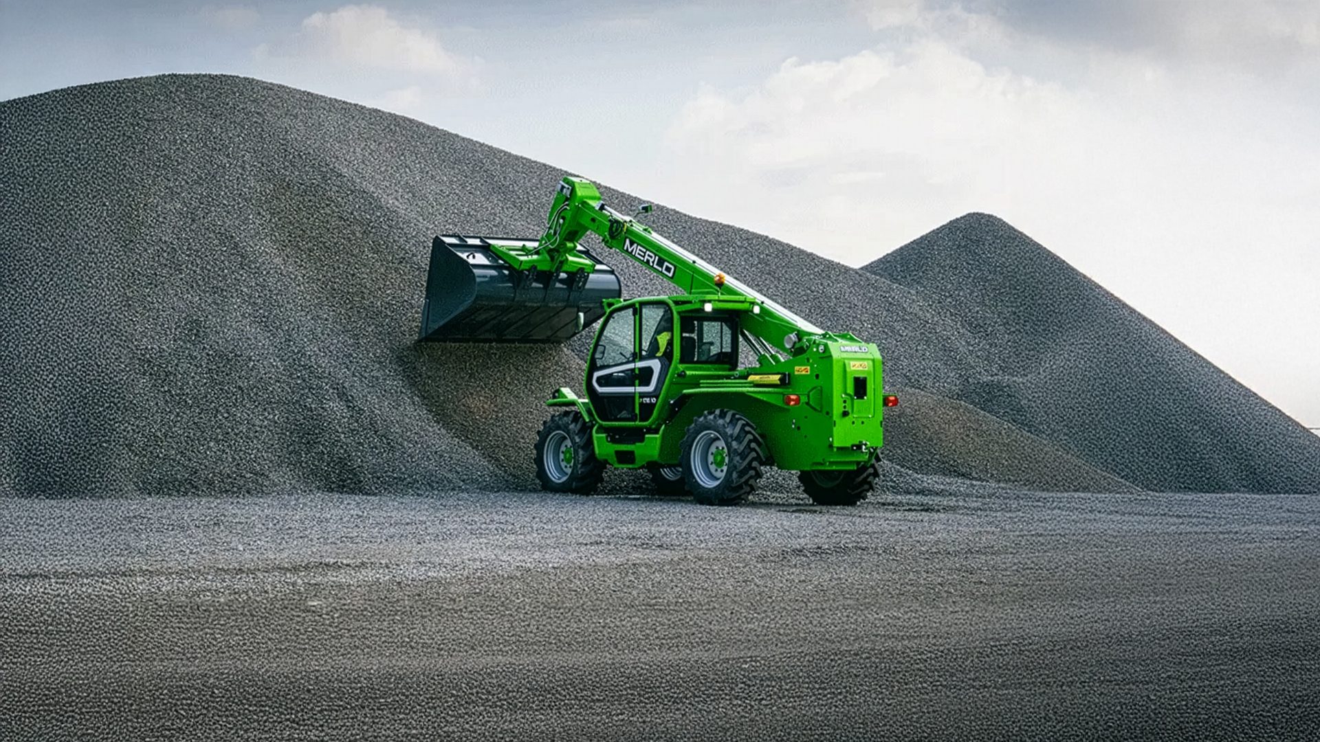 Green Merlo telehandler with its bucket in a large gravel pile.