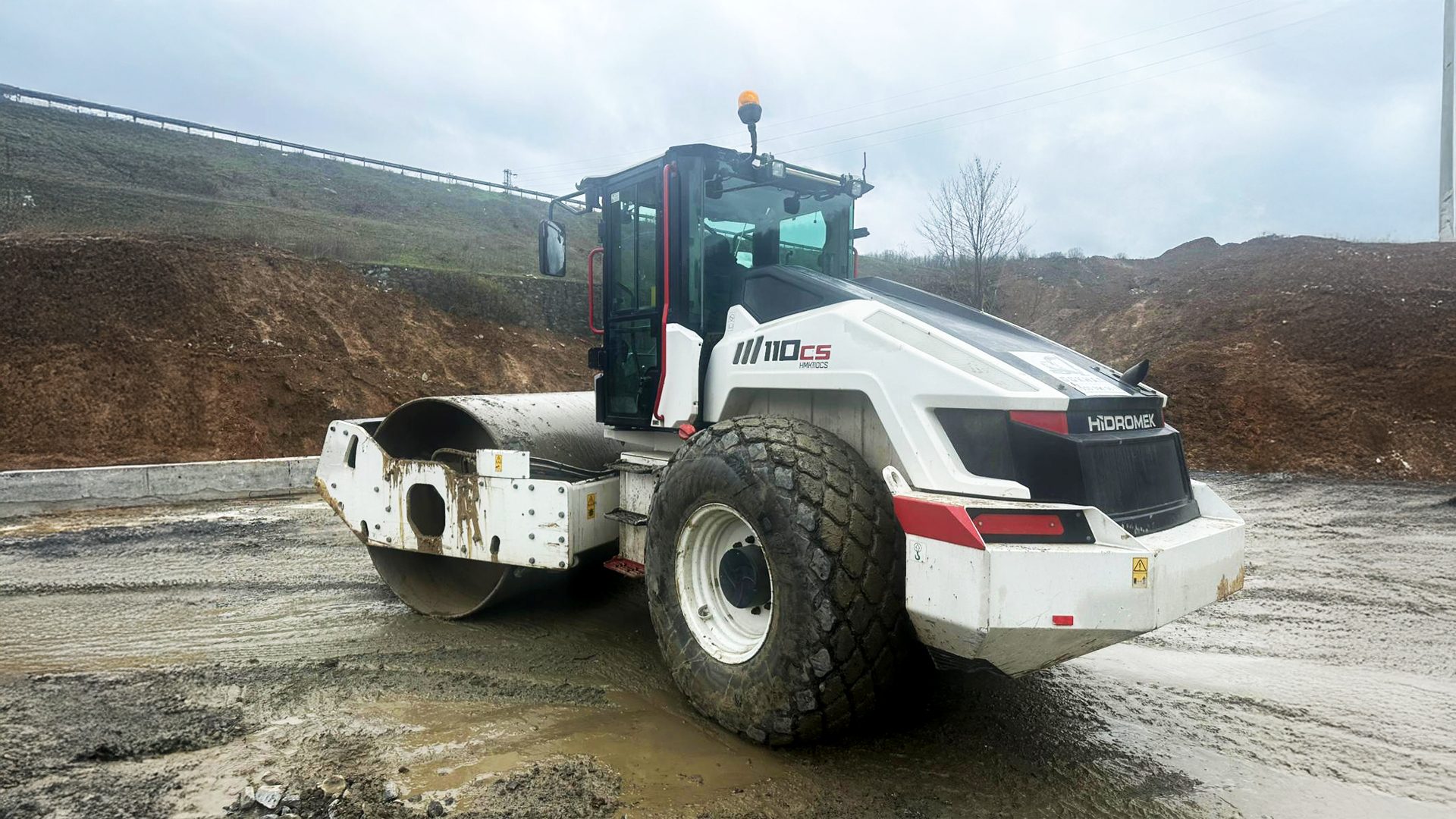 A white and black Hidromek 110CS road roller sits on muddy ground at a construction site.