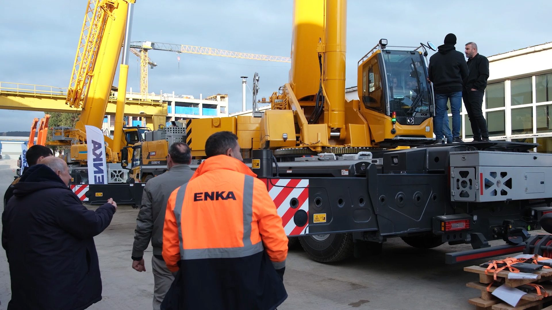 People inspect a large yellow mobile crane at an industrial site with other cranes and buildings.