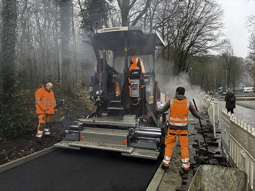 Workers in orange gear pave asphalt with a Dynapac machine, steam rises.