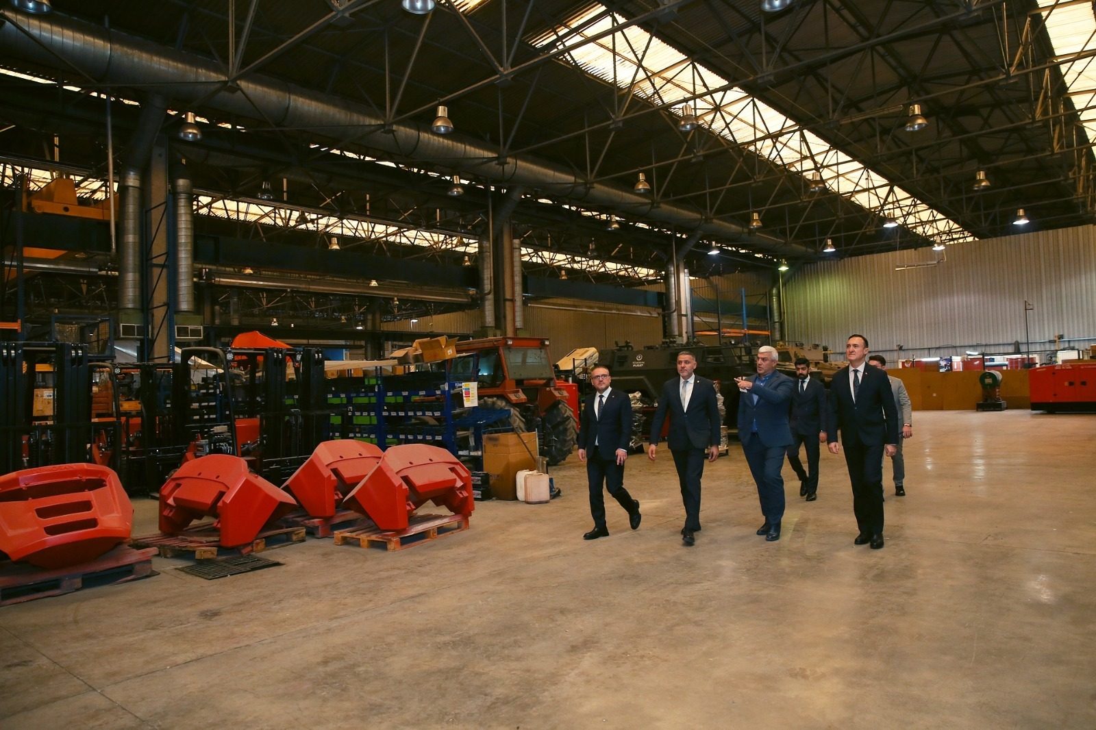 Men in suits walk through a large factory with machinery and red industrial parts.