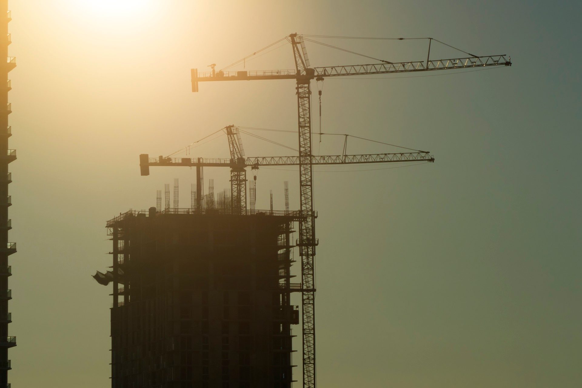 Silhouetted cranes and unfinished buildings at a construction site against a bright, hazy sunset sky.