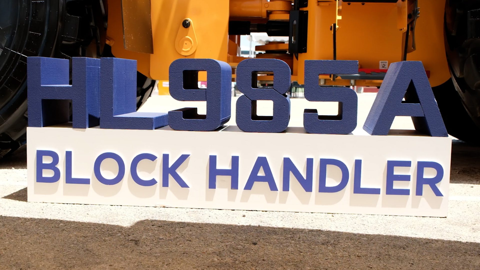 Blue 3D sign displays "HL985A BLOCK HANDLER" in front of yellow heavy machinery.