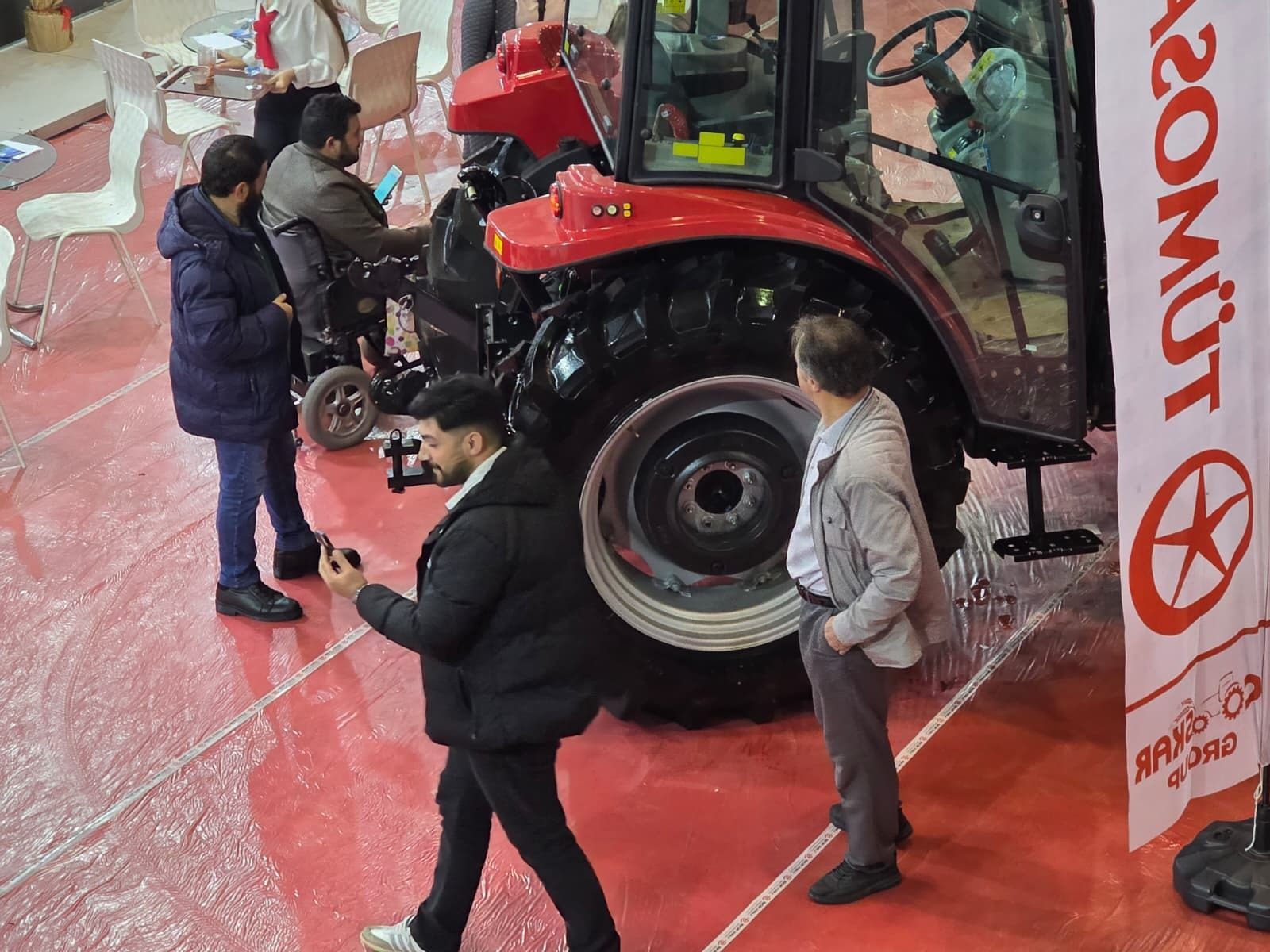 People gathered around a red tractor at an indoor exhibition, one person in a wheelchair.