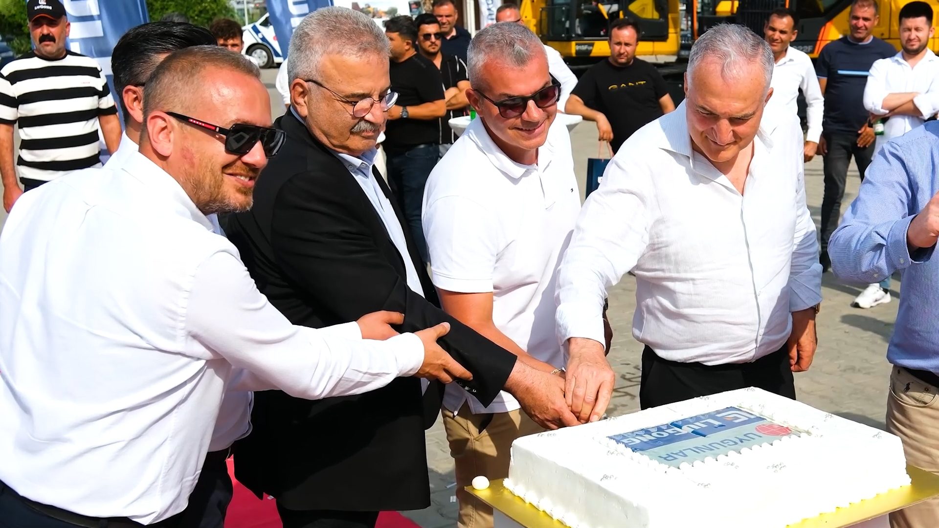 Four men cut a large white cake with a company logo at an outdoor event.