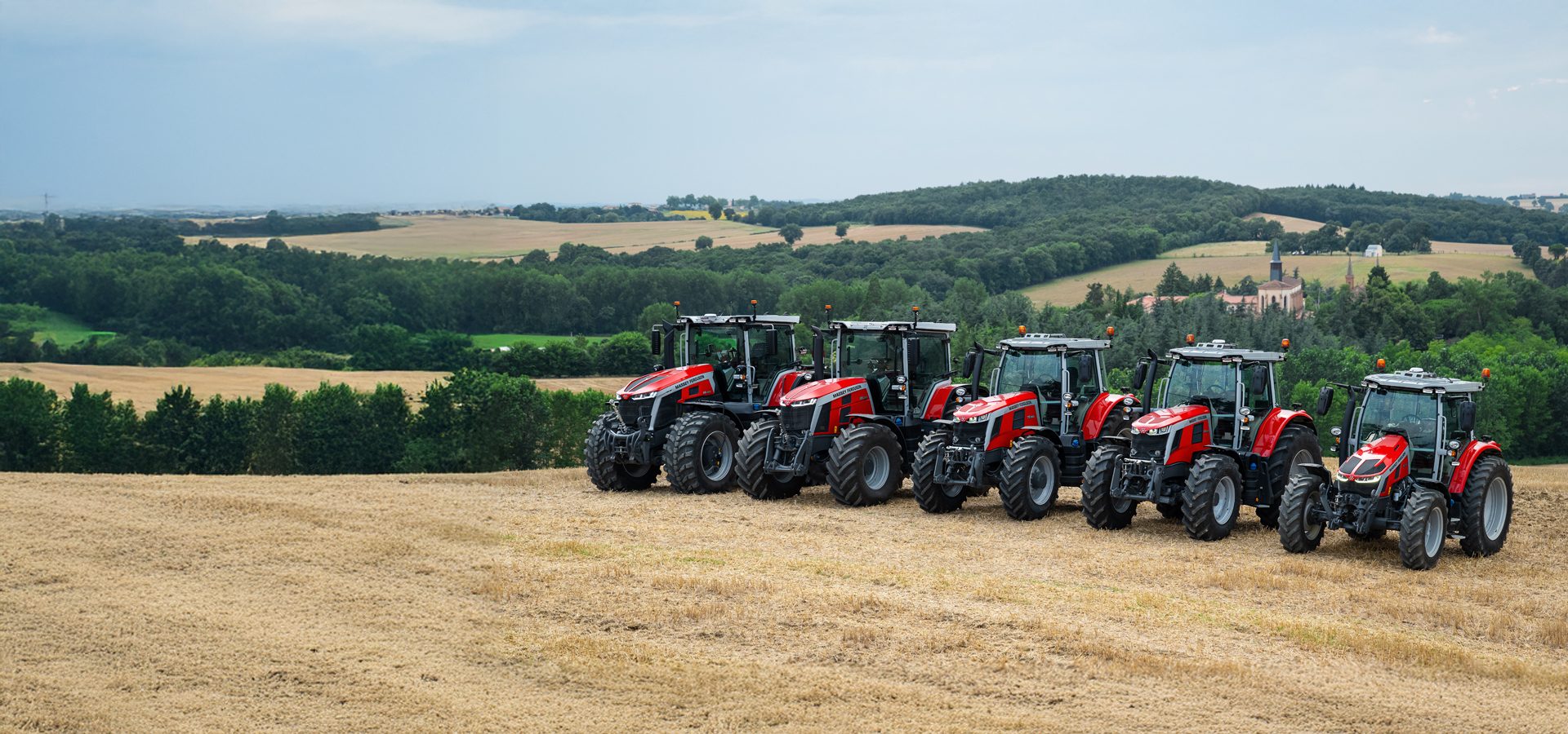 Six red tractors lined up in a golden field with rolling green hills in the background.