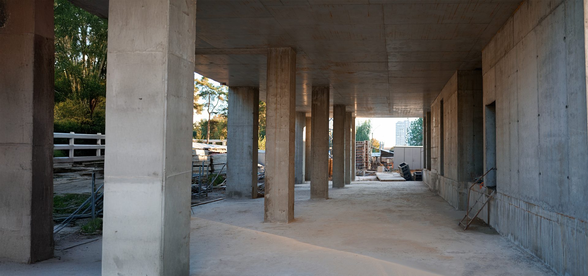 Unfinished concrete building with columns, open to greenery and a construction site.