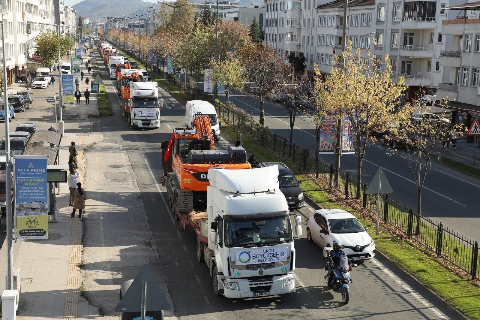 A convoy of trucks, some carrying excavators, drives down a city street with people on the sidewalk.