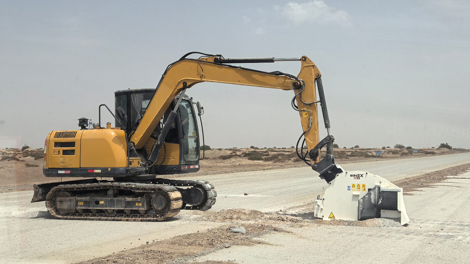 Yellow excavator with a road milling attachment on a dusty road in a desert landscape.