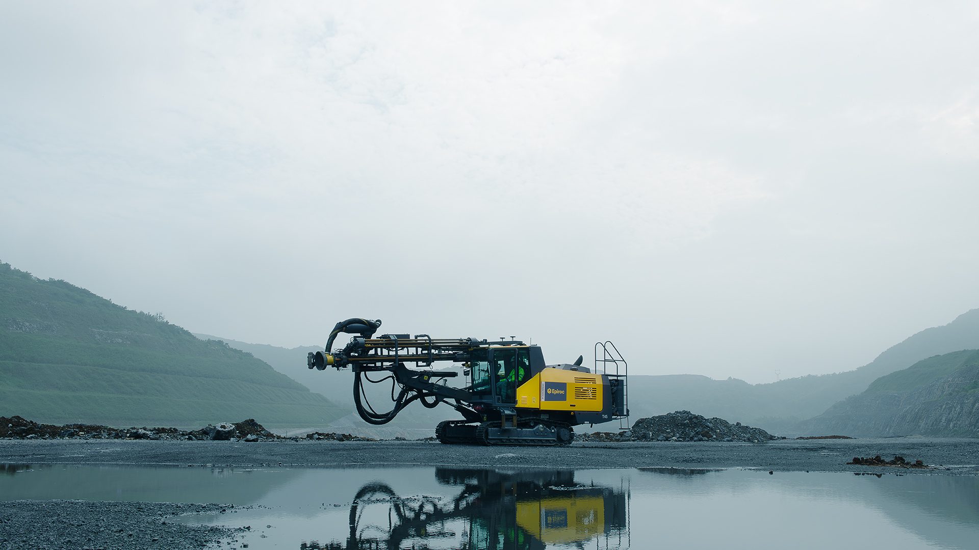 Epiroc T45 drilling rig on wet ground with hills and cloudy sky, reflected in water.