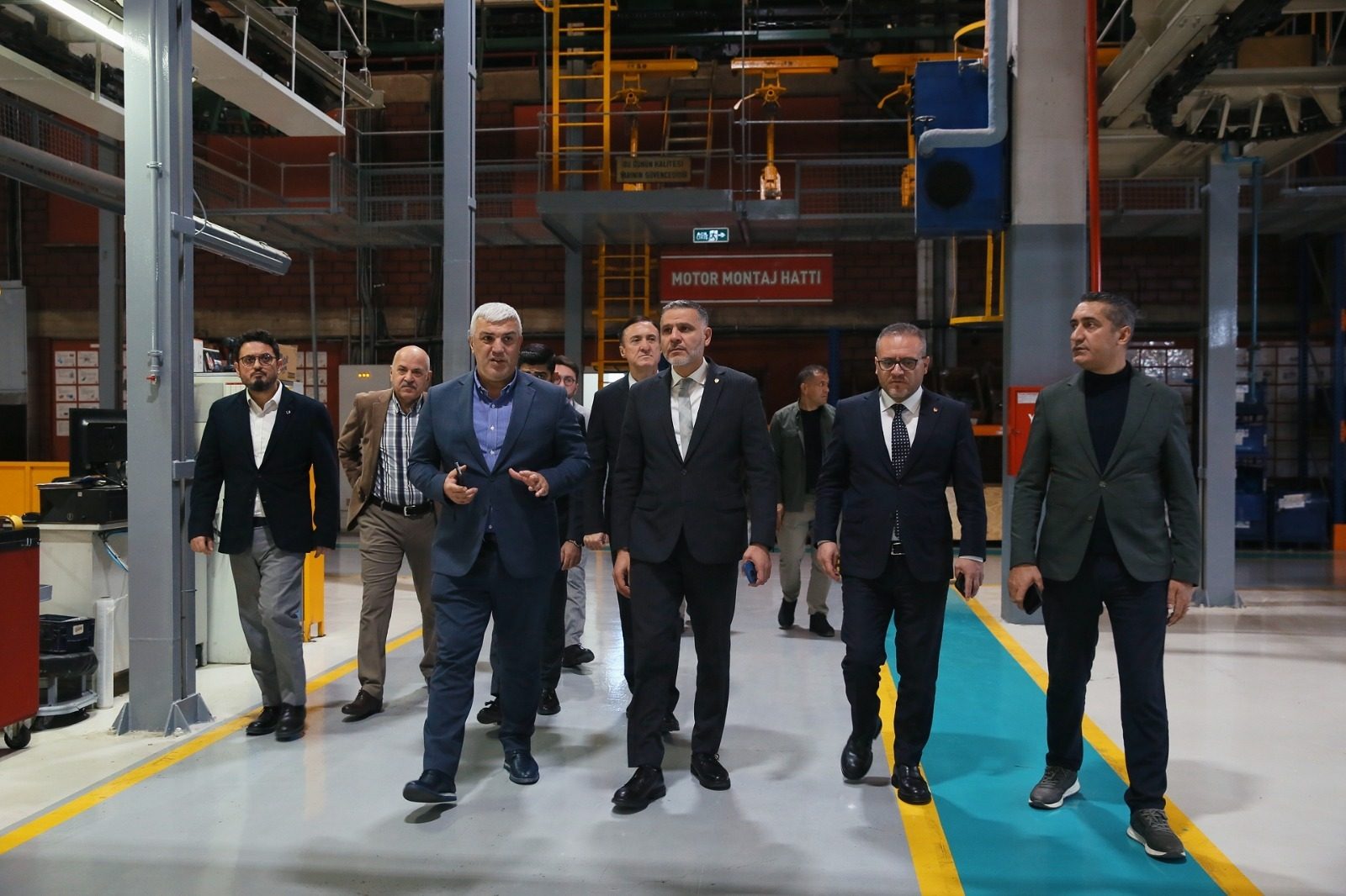 A group of men walk through a factory, past an "Engine Assembly Line" sign.