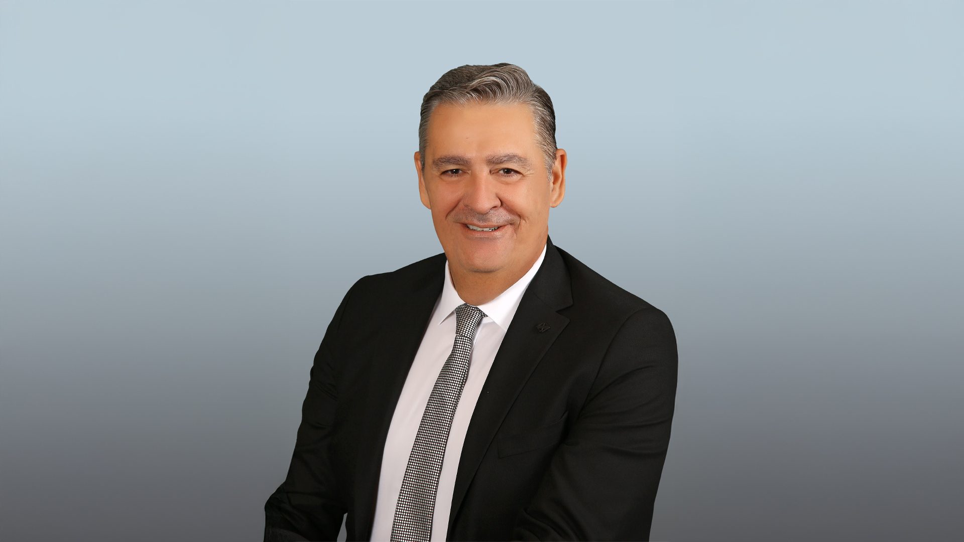 Smiling businessman in a black suit and patterned tie against a gradient background.