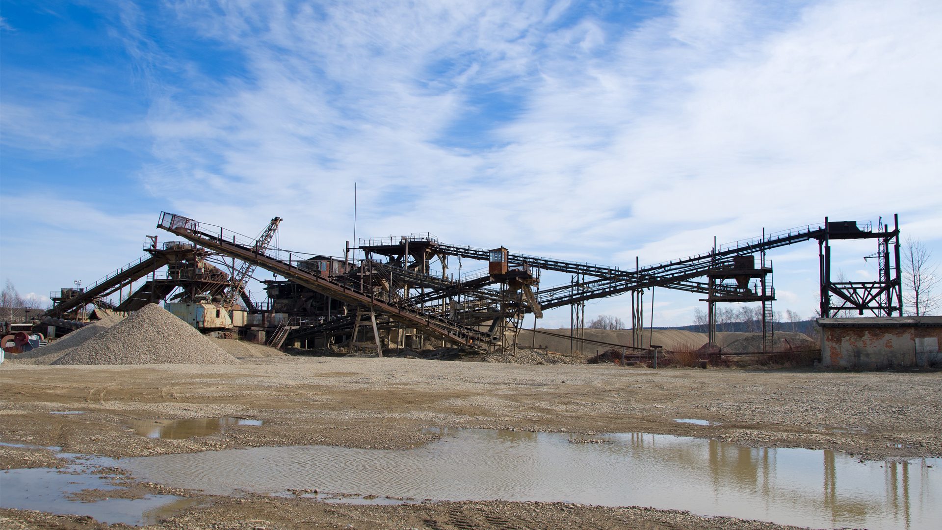 Industrial quarry with rusty conveyor belts, gravel piles, puddles, and a cloudy sky.