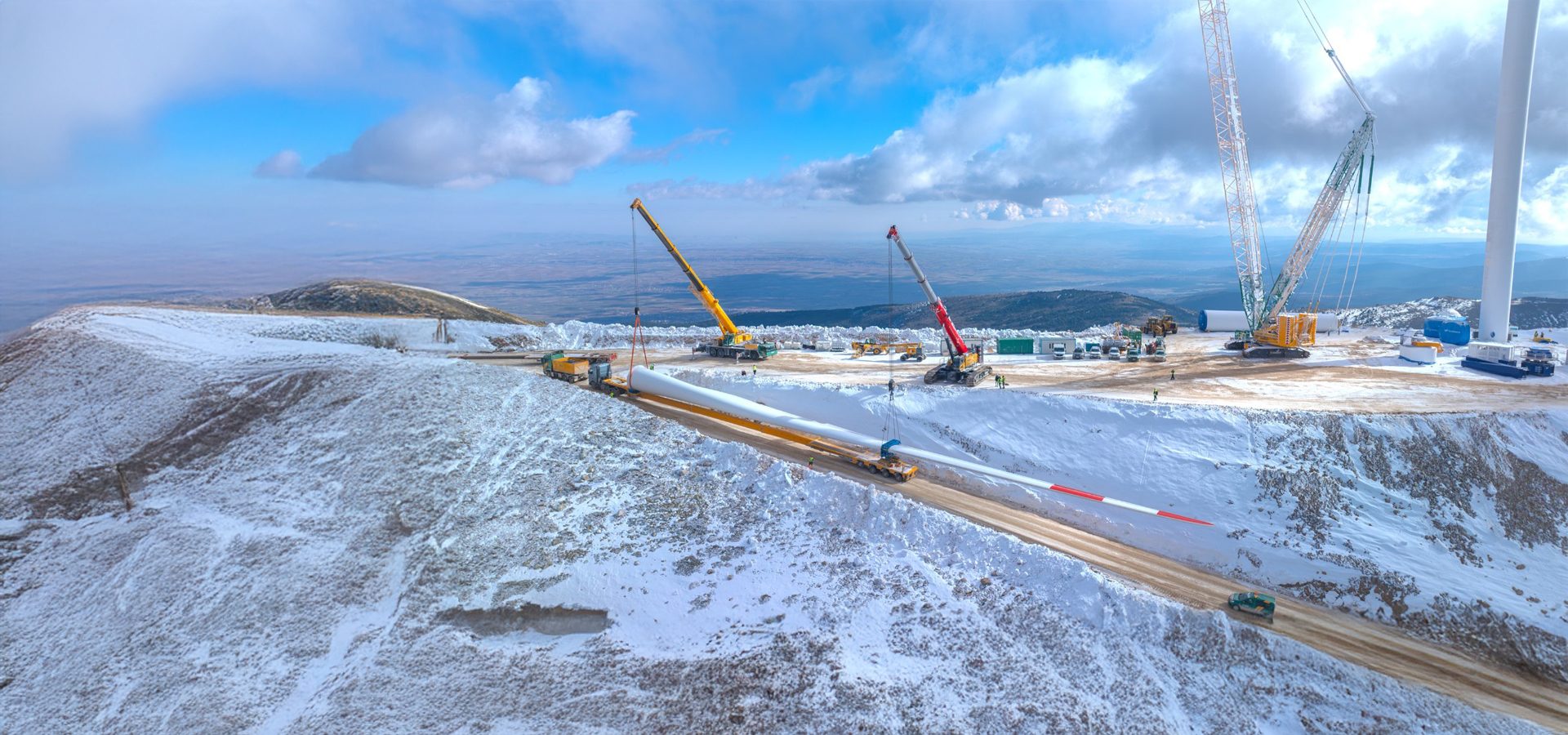 Wind turbine blade installation in a snowy mountain setting with cranes and workers.