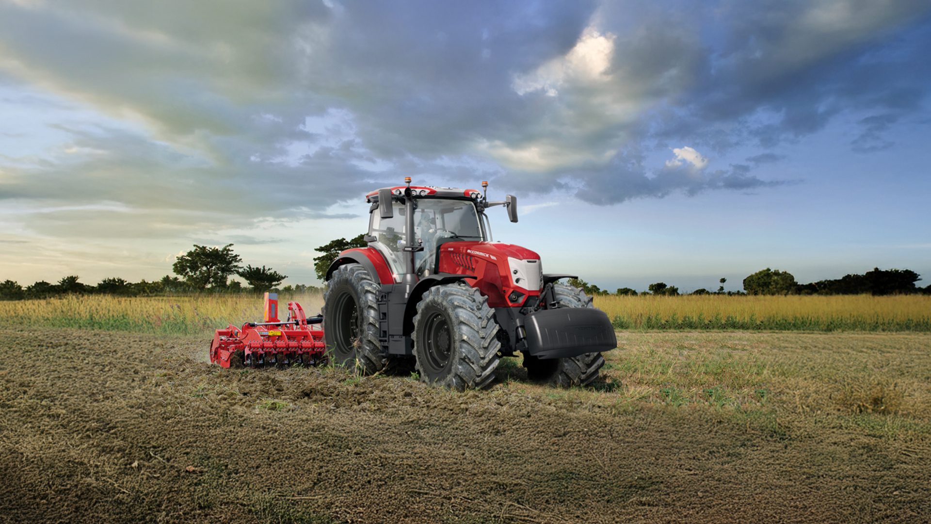 Red tractor with tillage implement in a field under a cloudy sky.