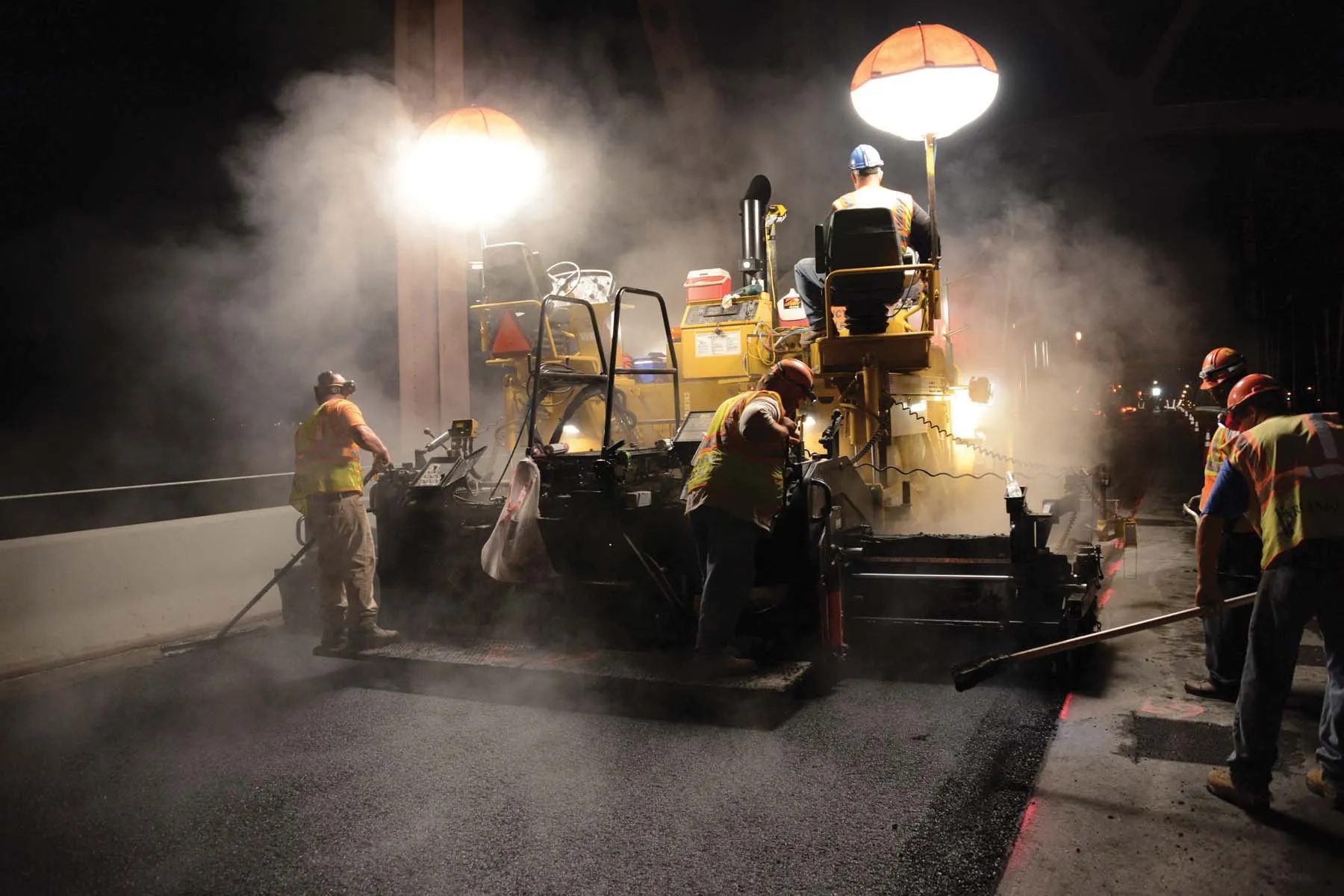 Night paving crew lays asphalt with heavy machinery and hand tools, illuminated by bright lights and steam.