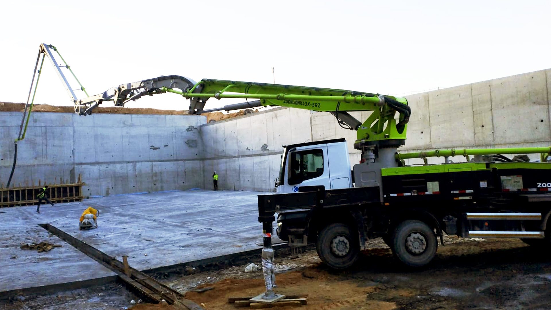 A concrete pump truck with a green boom extends over a construction site with concrete walls and workers.