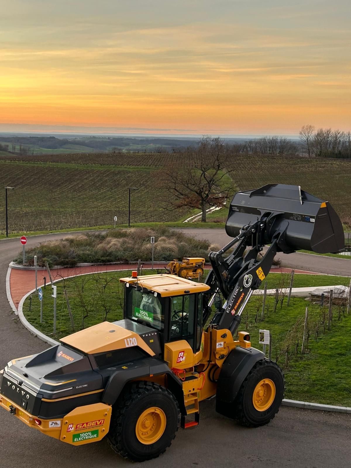 Volvo electric wheel loader with raised bucket in a vineyard landscape at sunset.