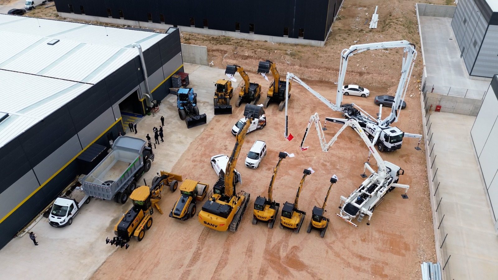 Aerial view of various construction vehicles and equipment displayed outdoors next to industrial buildings.