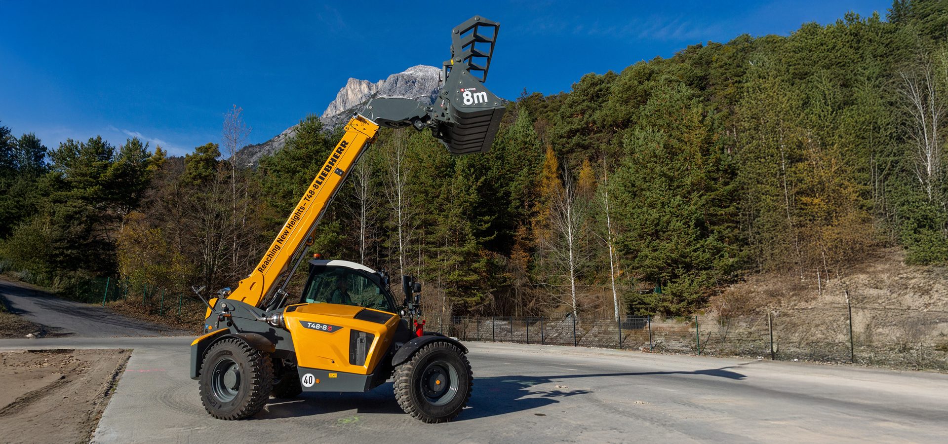 A yellow Liebherr telehandler with an extended arm and bucket, set against a backdrop of mountains and forests.