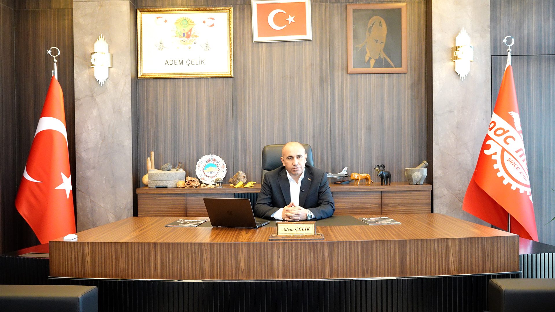 A man in a suit sits at a modern office desk, flanked by Turkish flags and framed art.