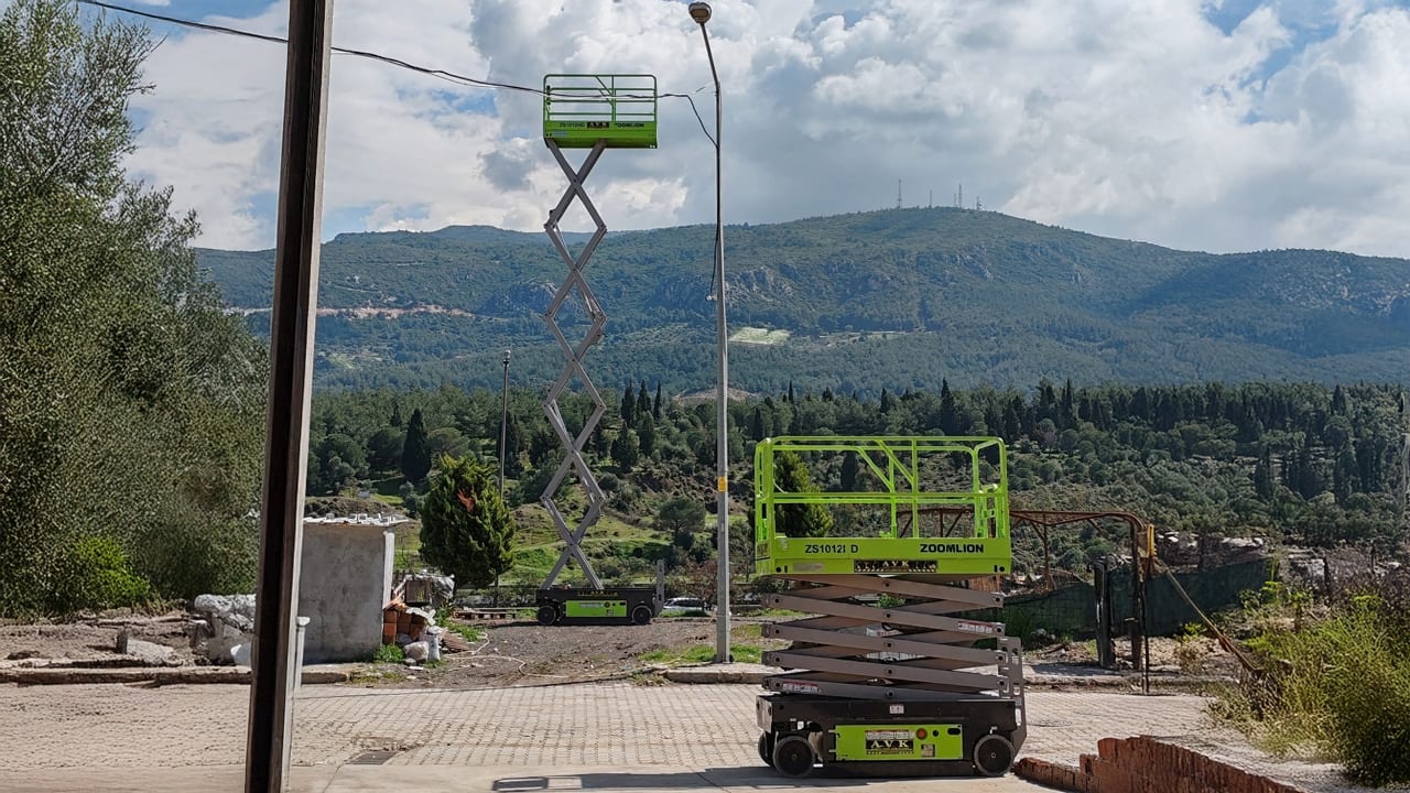Two green scissor lifts, one fully extended, in a mountainous landscape.