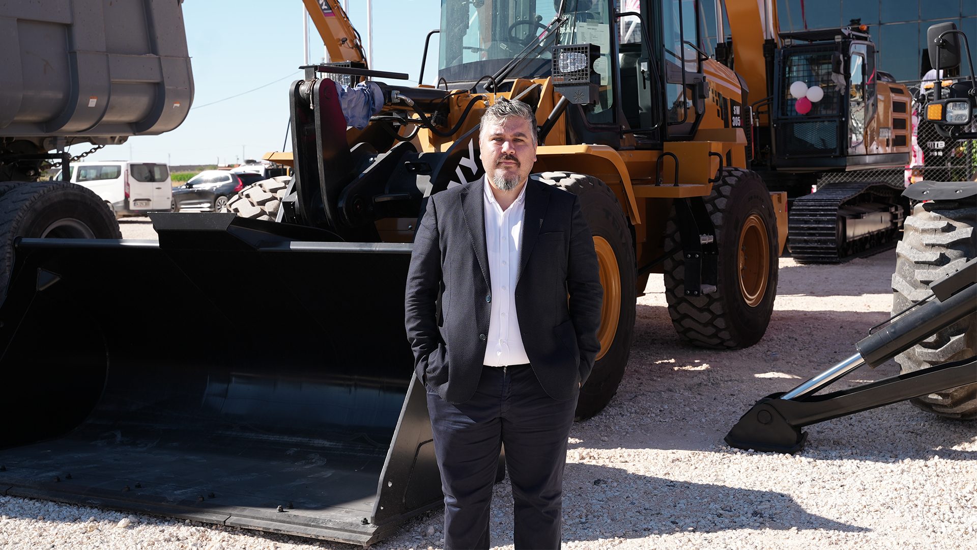 A man in a dark suit stands in front of yellow construction machinery, including a loader and excavator.