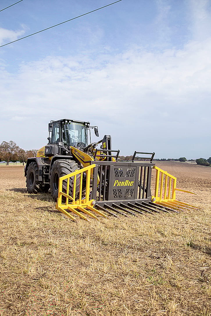 Yellow New Holland tractor with a Prollic agricultural fork attachment in a dry field under a cloudy sky.