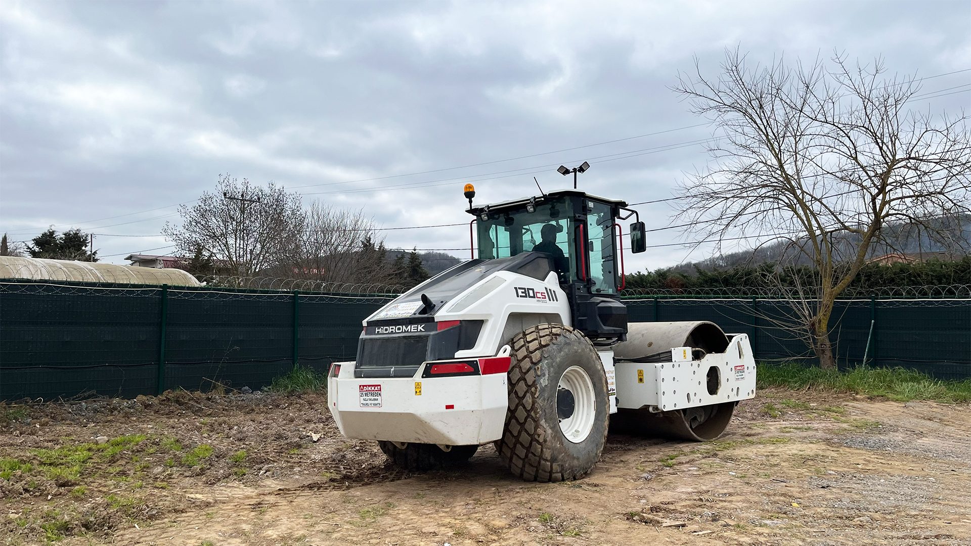 A white Hidromek 130CS III soil compactor parked on a dirt lot under a cloudy sky.