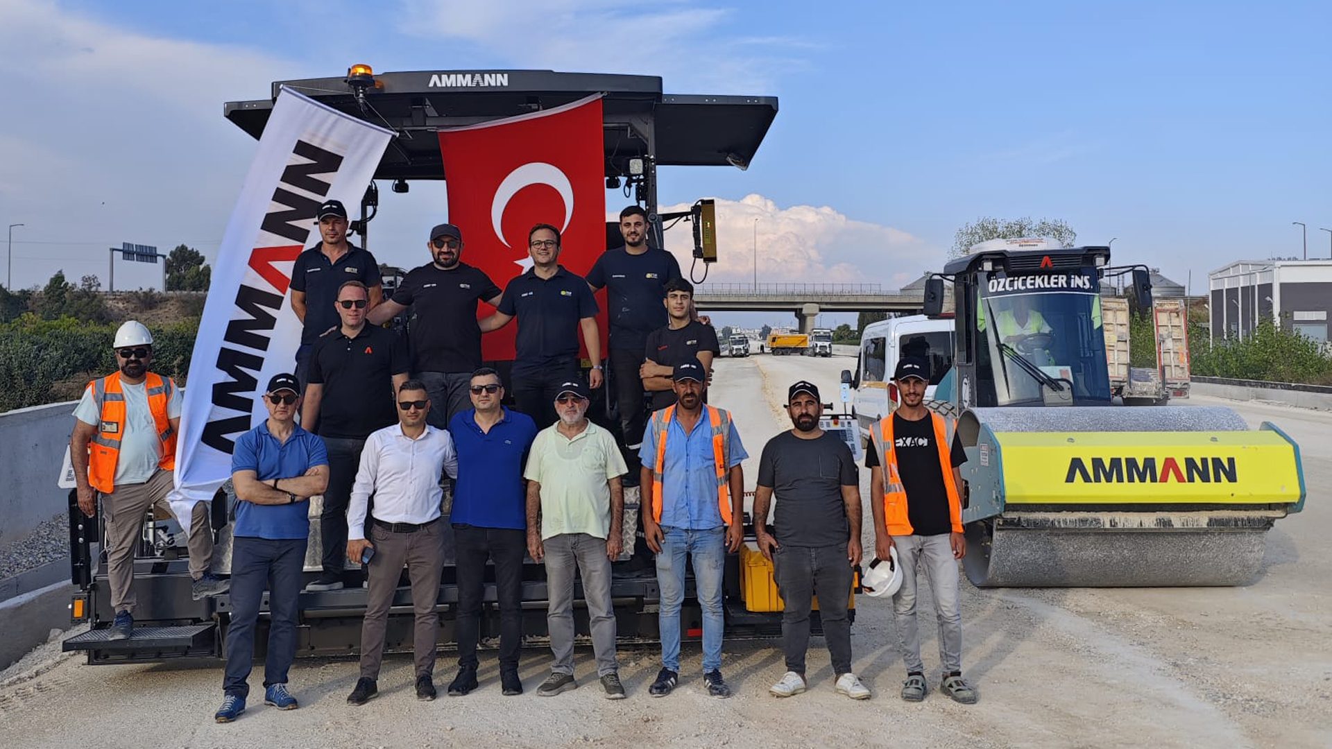 Construction workers pose with AMMANN road building machinery and a Turkish flag.