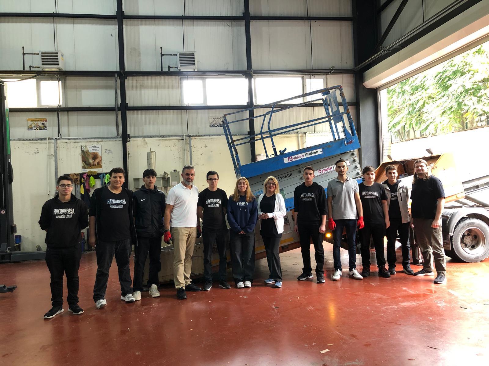A group of students and adults, some wearing "HAYDARPASA" shirts, pose in a workshop with machinery.