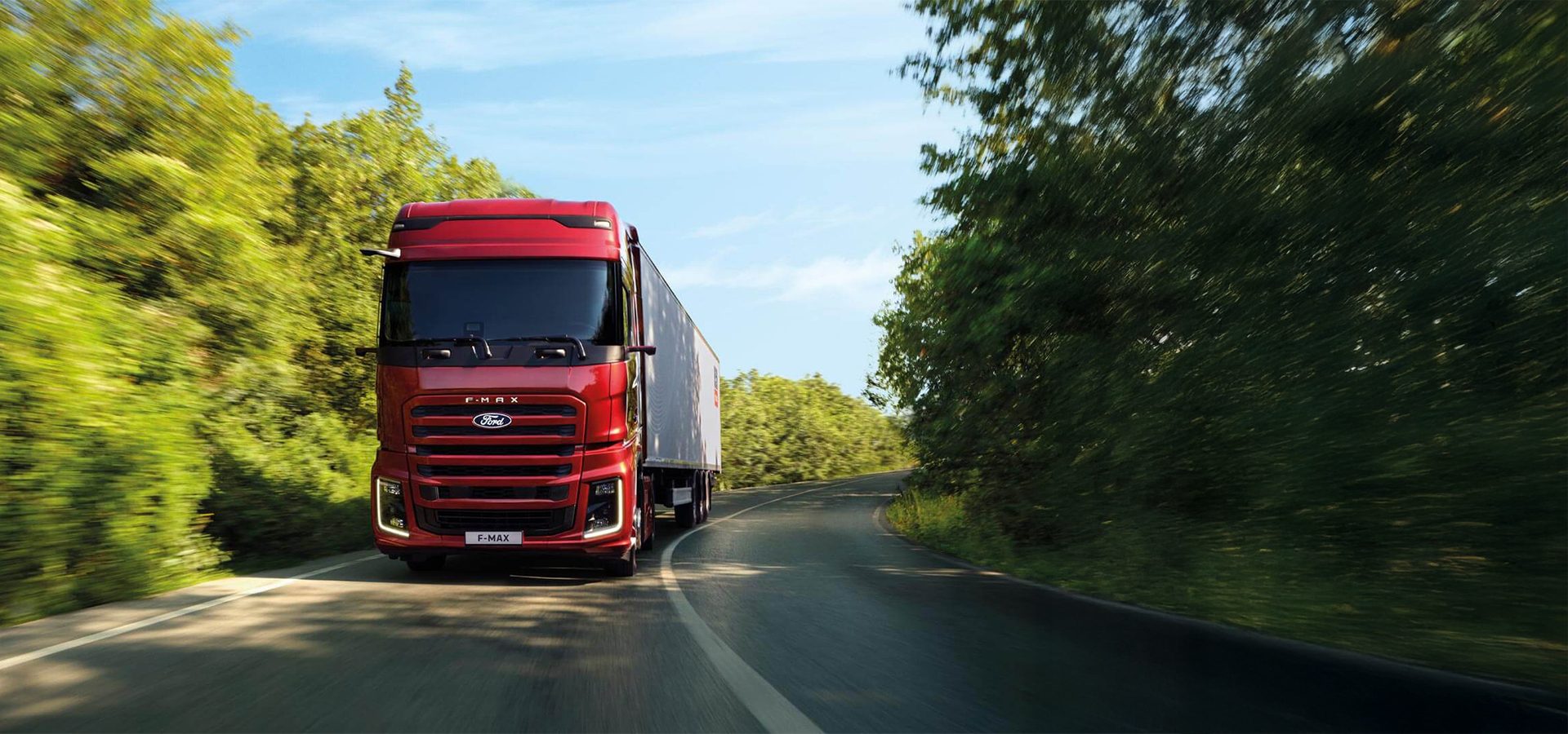 Red Ford F-Max truck with a trailer driving on a winding road through lush green trees under a blue sky.