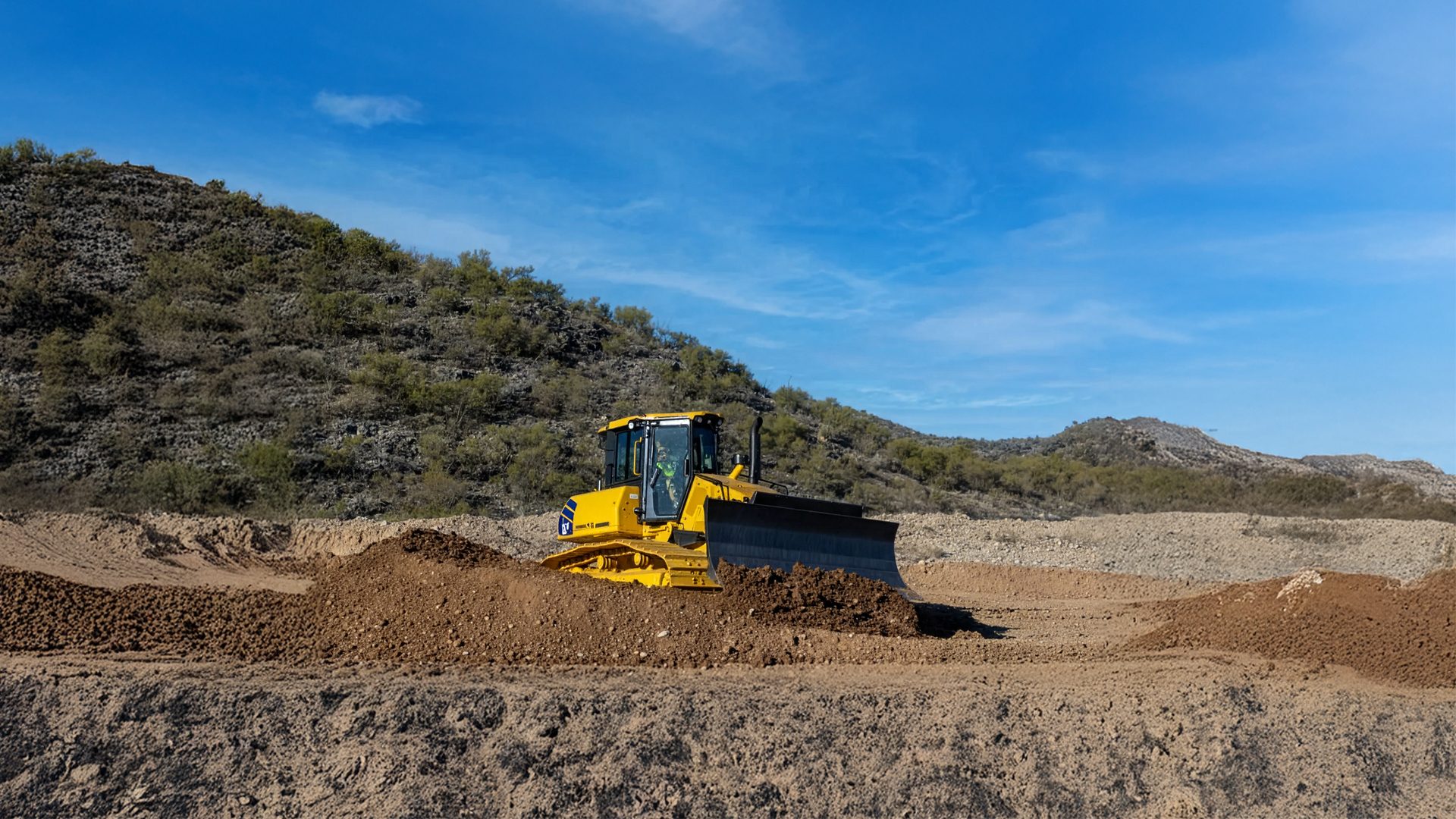 A yellow bulldozer moves dirt on a construction site with arid hills and a blue sky in the background.