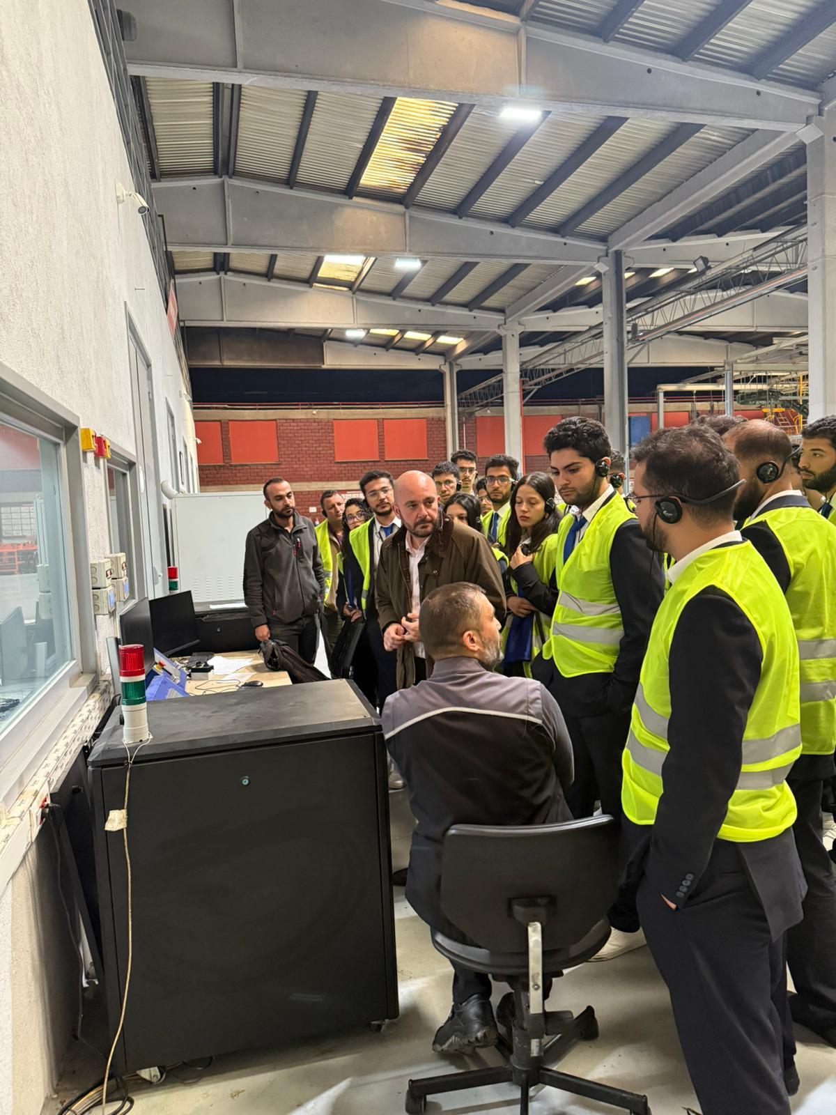 A group of visitors, many in safety vests and headphones, observe an industrial control station.
