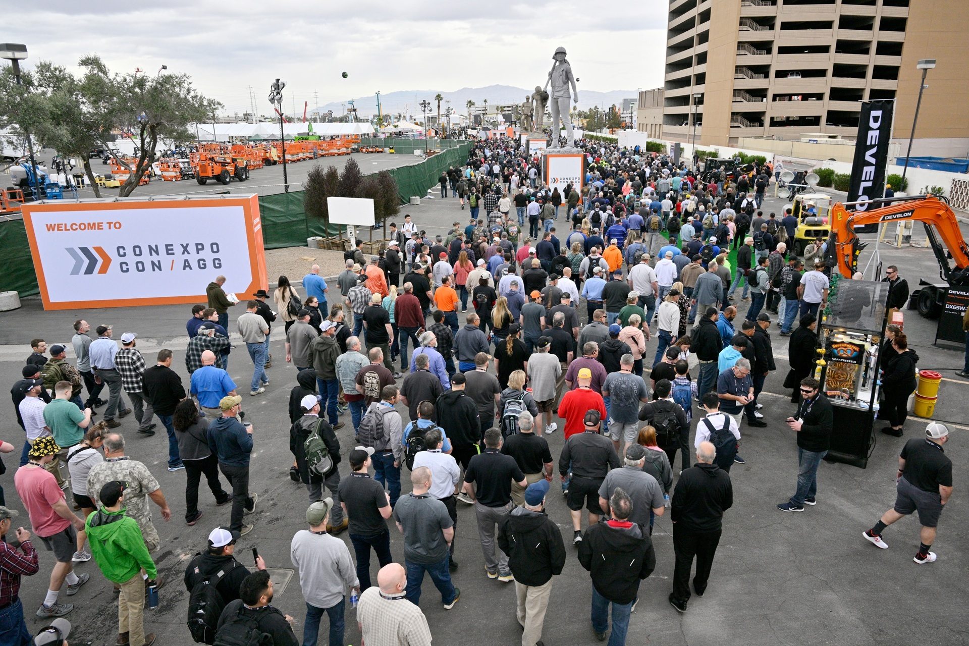 A large crowd gathers outdoors at CONEXPO CON/AGG, with construction equipment and a large statue.