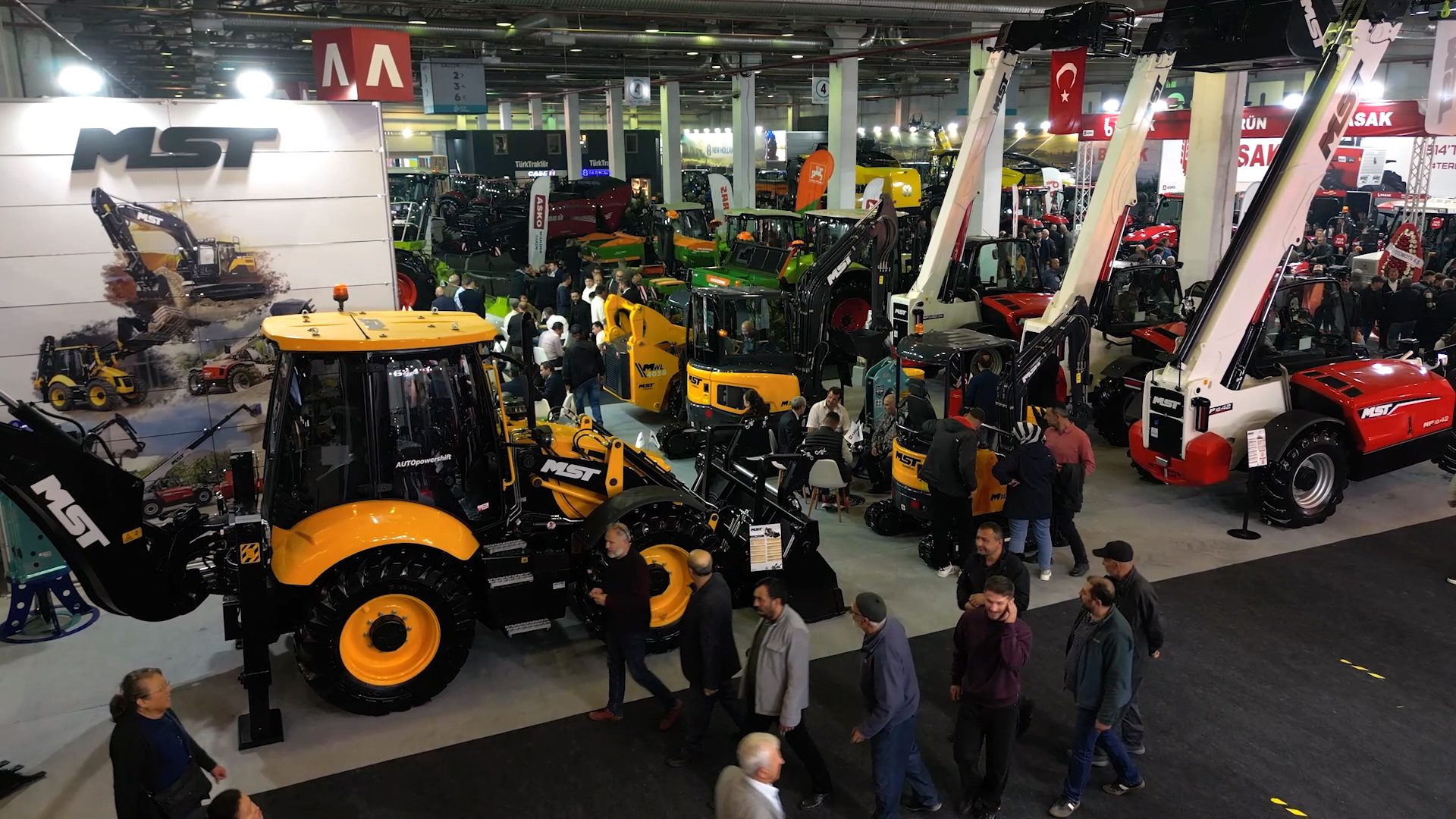 Crowded exhibition hall displaying MST construction machines: yellow backhoes and red telehandlers.