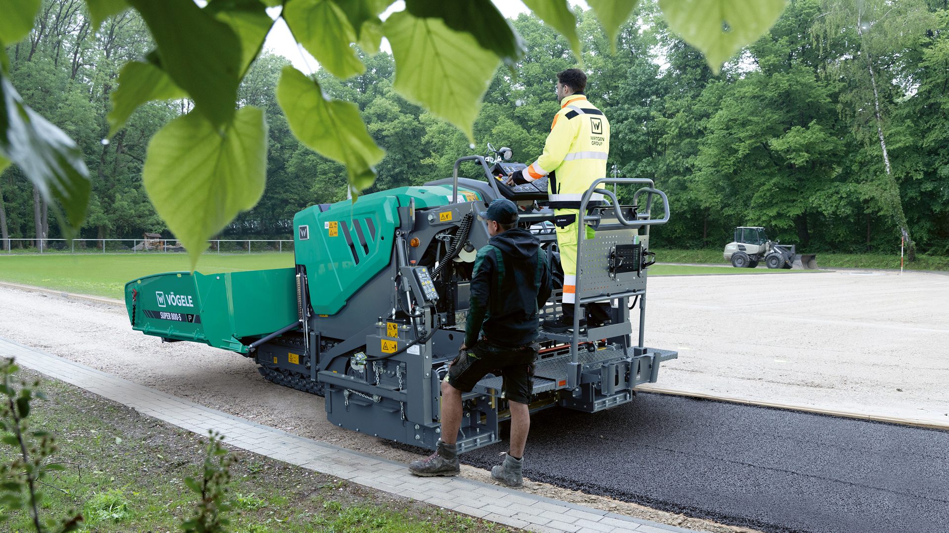 Two workers operate a green Vögele asphalt paver, laying fresh asphalt by a grassy field.