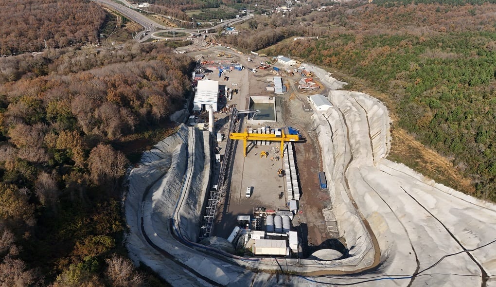 Aerial view of a tunnel construction site with crane and equipment in a wooded area, road visible.