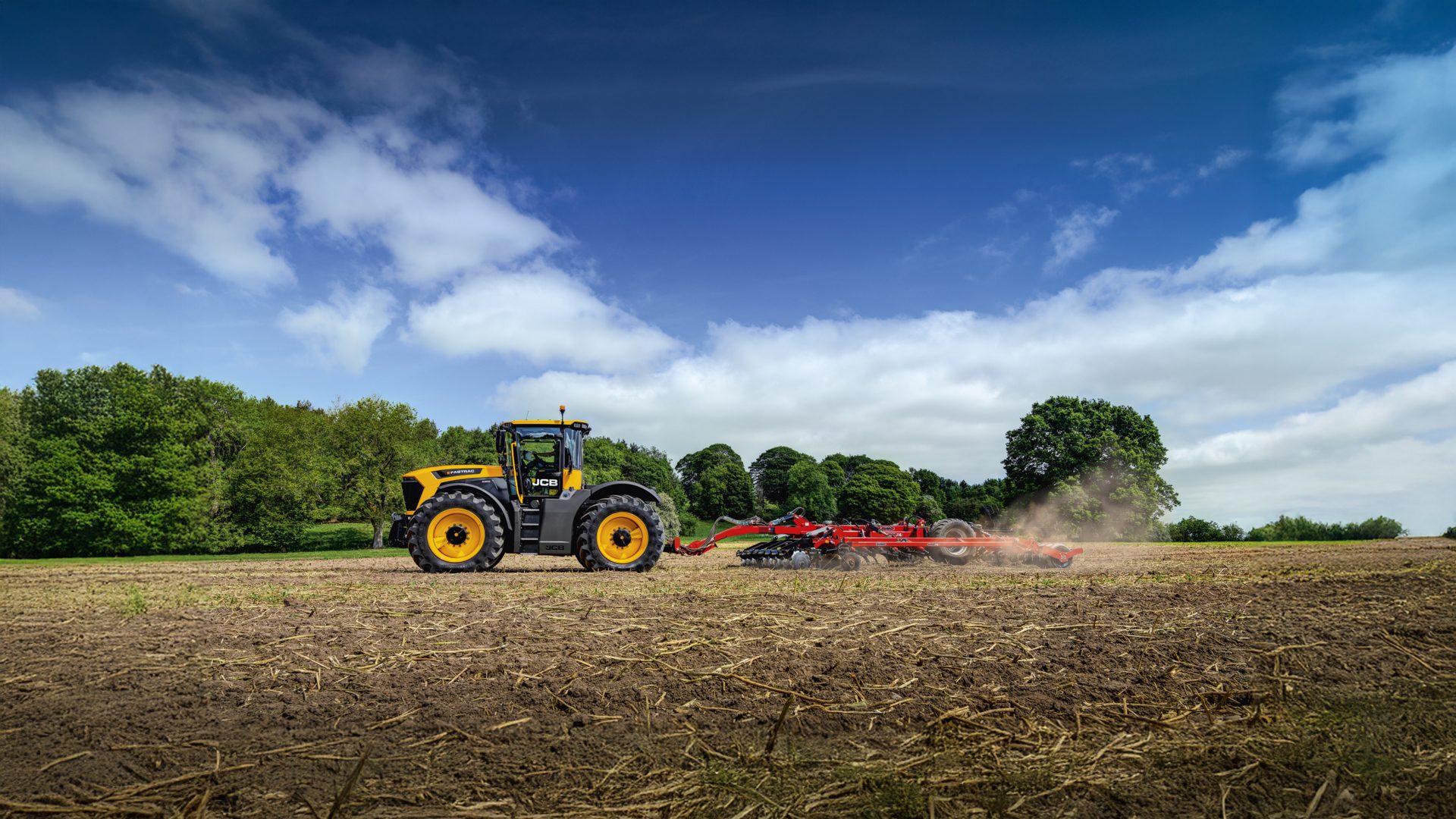 Yellow JCB tractor cultivating a field under a blue sky, kicking up dust.
